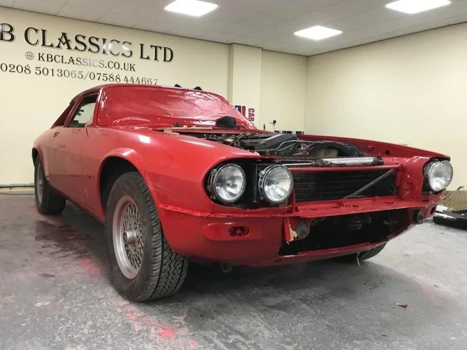 A red car with its front bumper removed, revealing internal parts, parked inside a garage with a white wall. The car appears to be a classic model and is missing its headlights and grille.