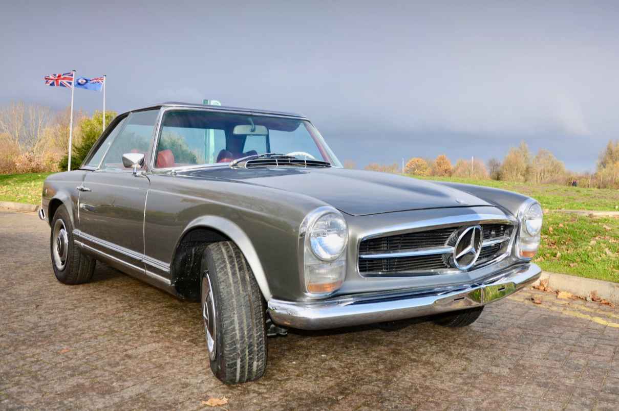 A vintage Mercedes-Benz car parked on a paved surface with flags in the background under a cloudy sky.