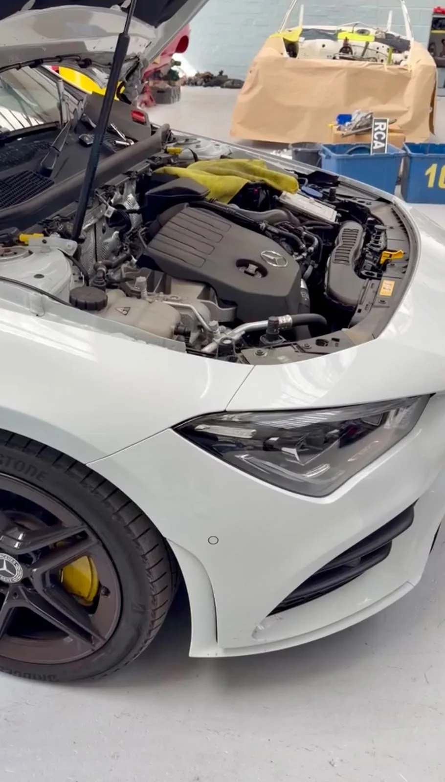 A white sports car with the hood open, showing the engine inside a garage or workshop with tools and a boat in the background.