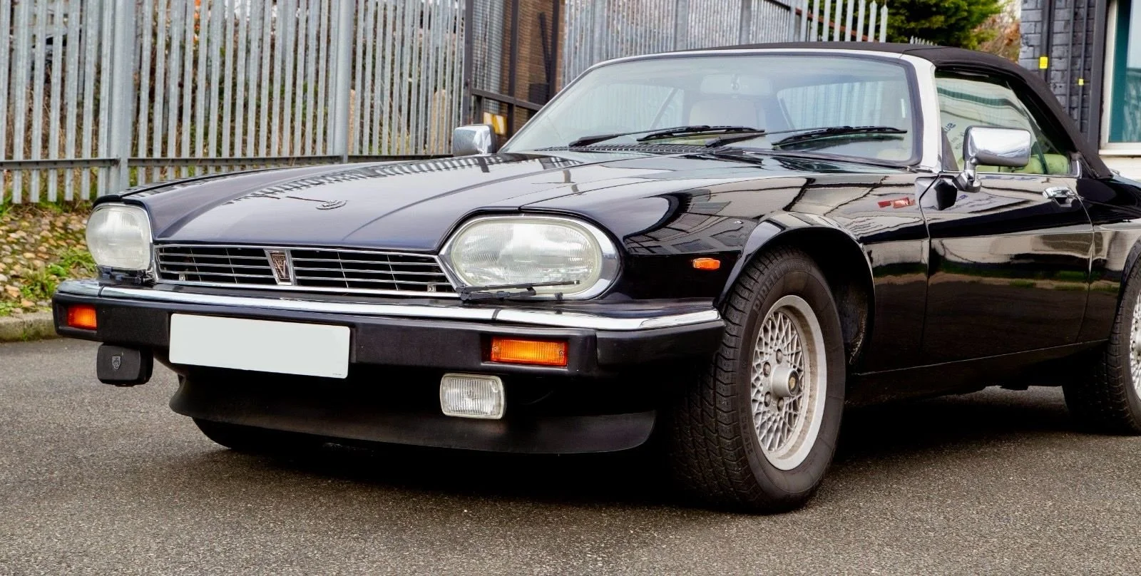 A black vintage convertible car parked on a gravel surface with green trees and a cloudy sky in the background.