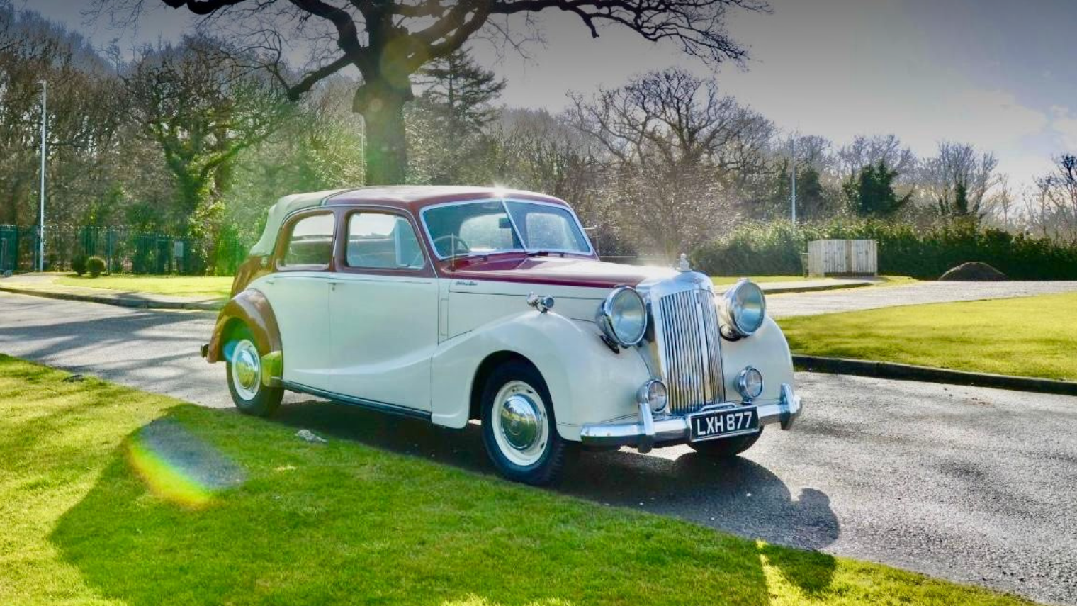 A vintage cream-colored car with maroon accents parked on a street near a grassy area, with trees and a sunny sky in the background.