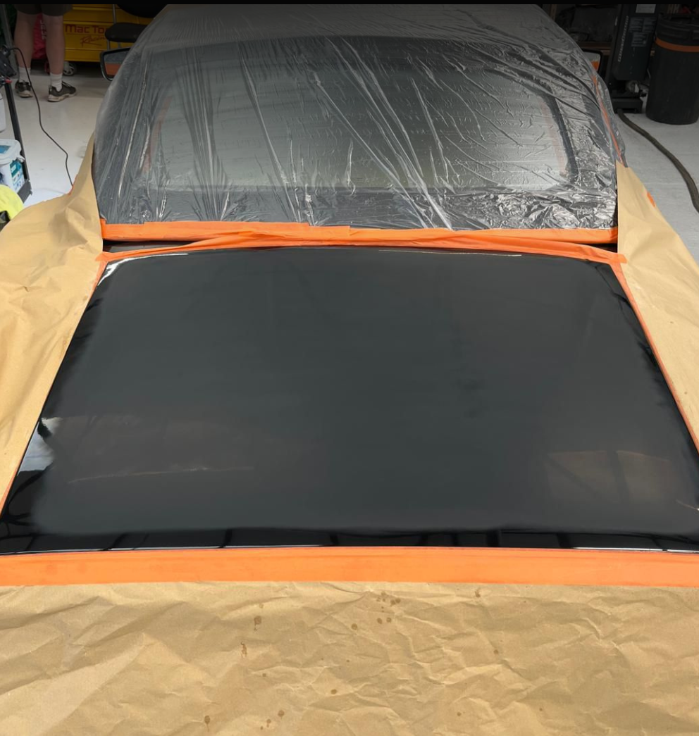 The image shows a car in the process of being painted or detailed. The hood of the car is freshly painted in a glossy black finish, and the surrounding areas are covered with masking paper and plastic sheeting to protect them from paint overspray.