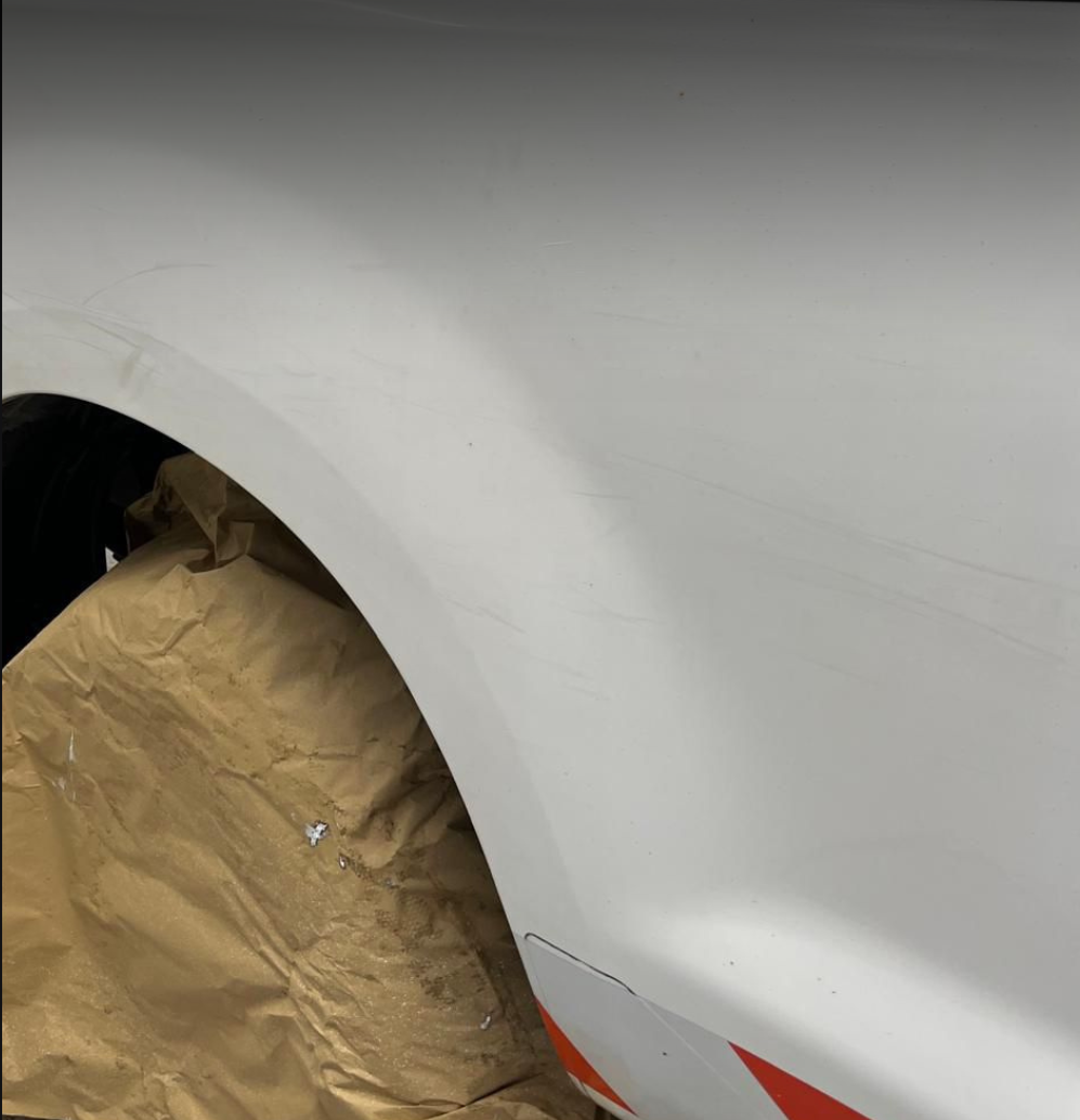 Close-up of a white car with scratches on the front fender, partially covered with brown paper, possibly in a repair shop.