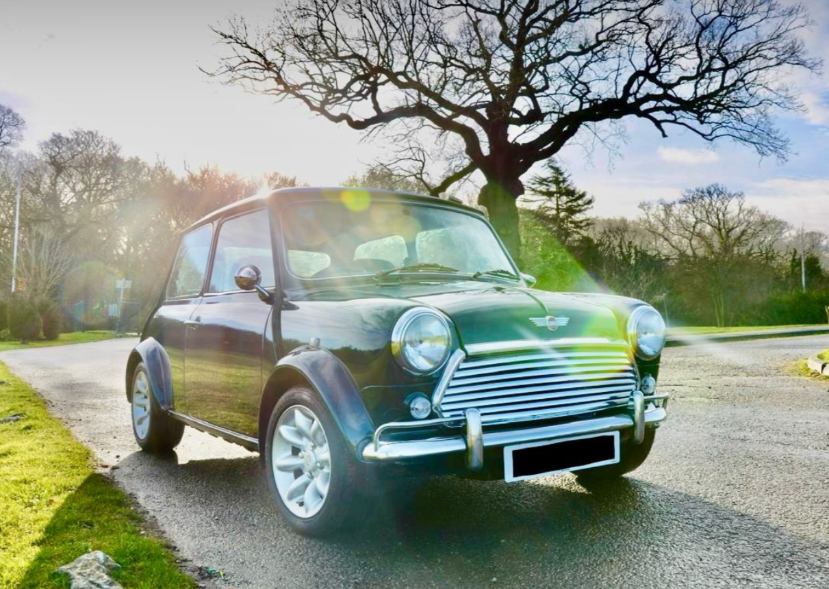A dark green Mini Cooper car parked on a paved road beside a grassy area with trees, sunlight, and a partly cloudy sky in the background.