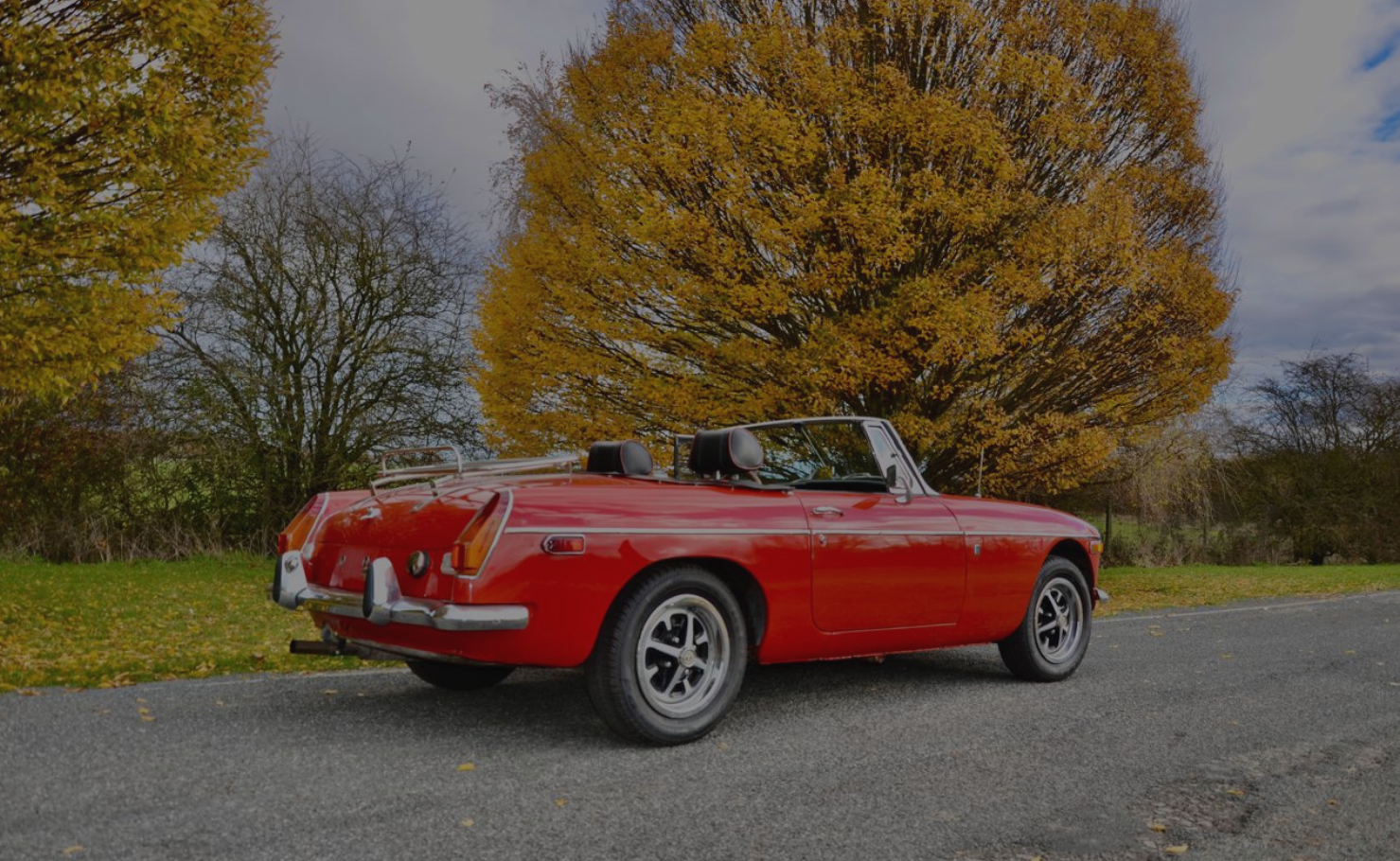 A red vintage convertible car parked on side of the road with autumn trees with yellow and orange leaves in background.