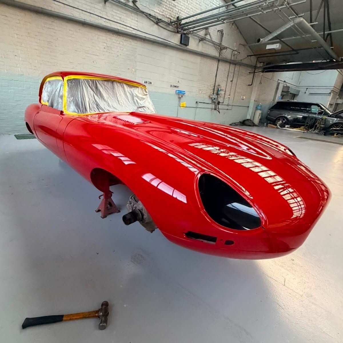 A vintage red race car body shell on jack stands inside a workshop, with a hammer on the floor nearby and other cars in the background.