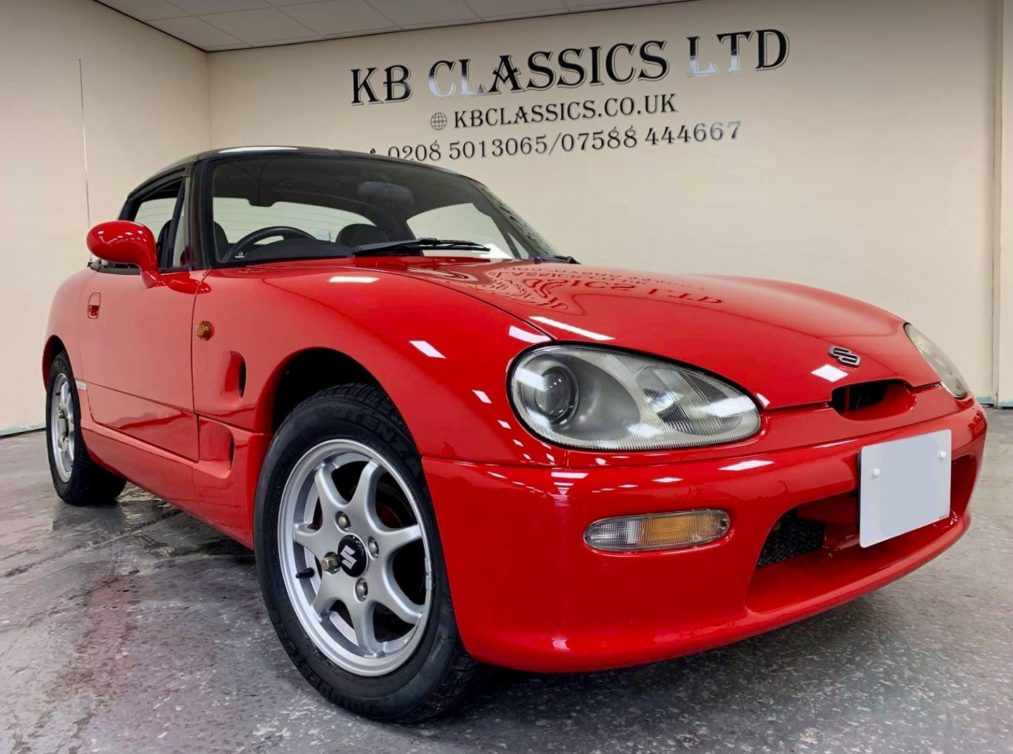 Red Suzuki sports car with black roof parked indoors in front of a wall with "KB Classics LTD" sign and contact information.