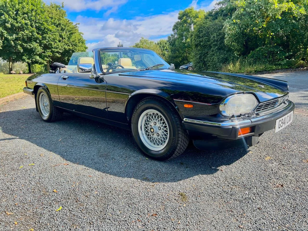 Black vintage convertible sports car parked on a gravel surface with green trees in the background and a partly cloudy sky.