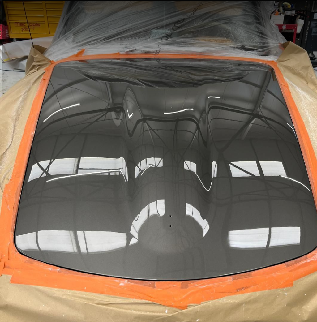 A car hood that has been painted with a glossy black finish, reflecting the ceiling of a workshop or garage, with protective masking tape and paper around the edges.