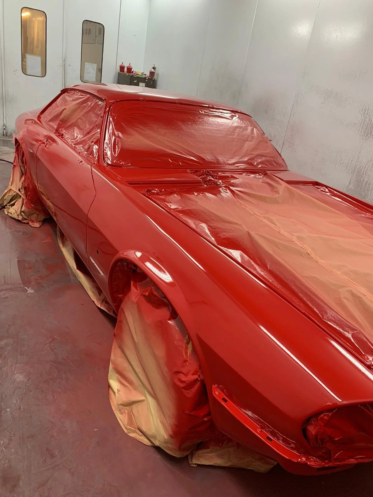 A red car is being painted in a spray booth, with plastic covering parts to protect them from paint overspray.