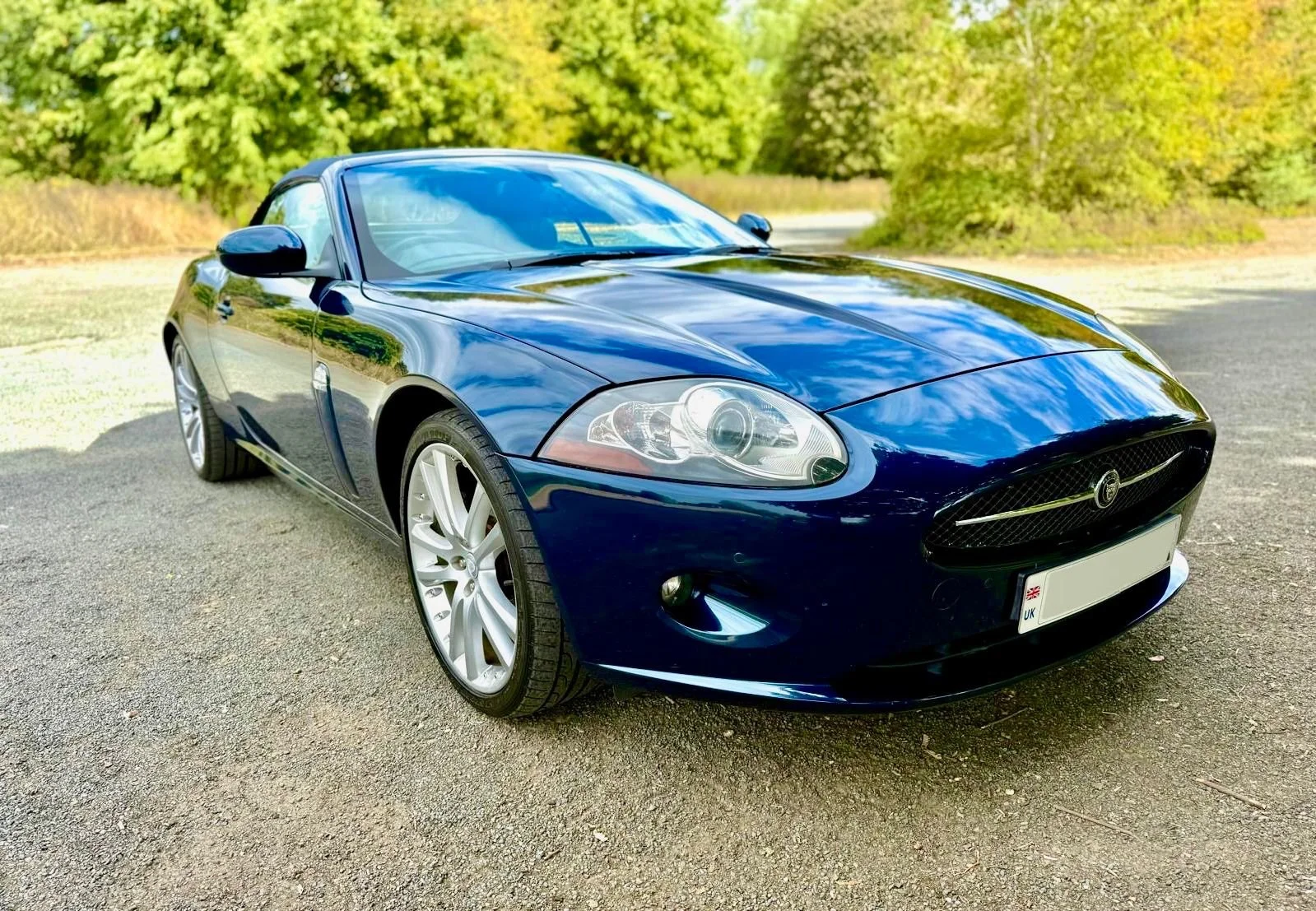A shiny black convertible sports car parked on a gravel driveway with lush green trees in the background.