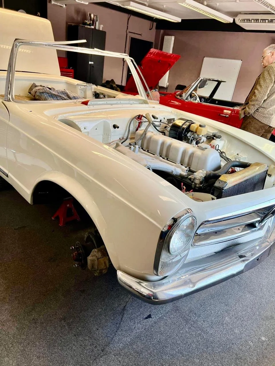 A vintage white convertible car with its hood open, showing the engine. The car is on a floor with no wheels, resting on red jack stands.