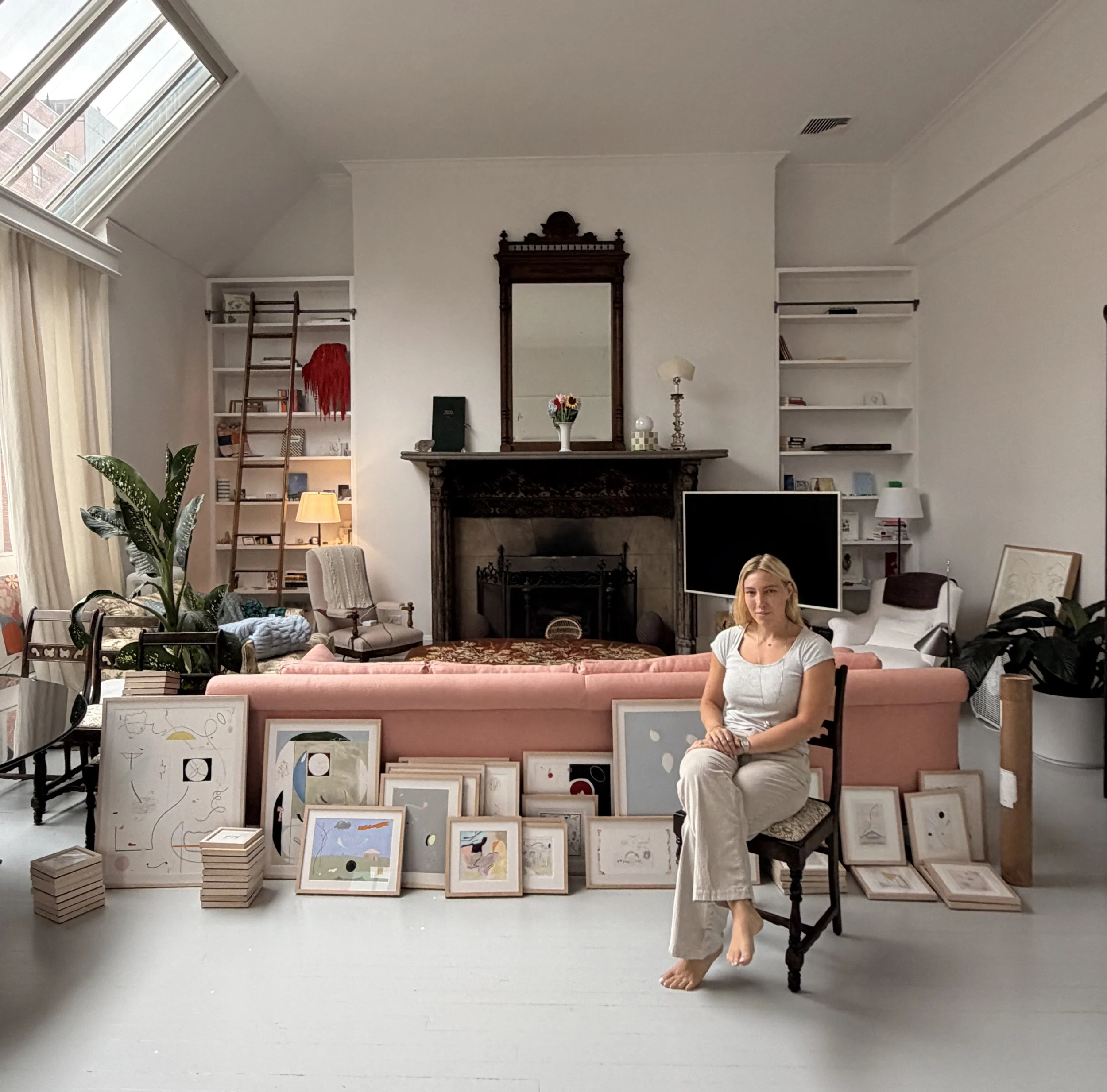 Interior of an art gallery or bookstore with white walls and high ceiling, featuring a variety of framed artwork displayed on racks and leaning against a pink sofa. There are paintings of abstract and minimalist styles, a woman dressed in white sitting on a chair, and various decorative items including books, plants, and lamps. Natural light floods the space through a large skylight and window with curtains.