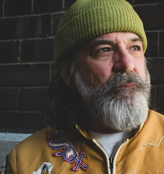 Close-up of Joe Pernice wearing a green knit beanie and a tan jacket, standing against a dark brick wall.