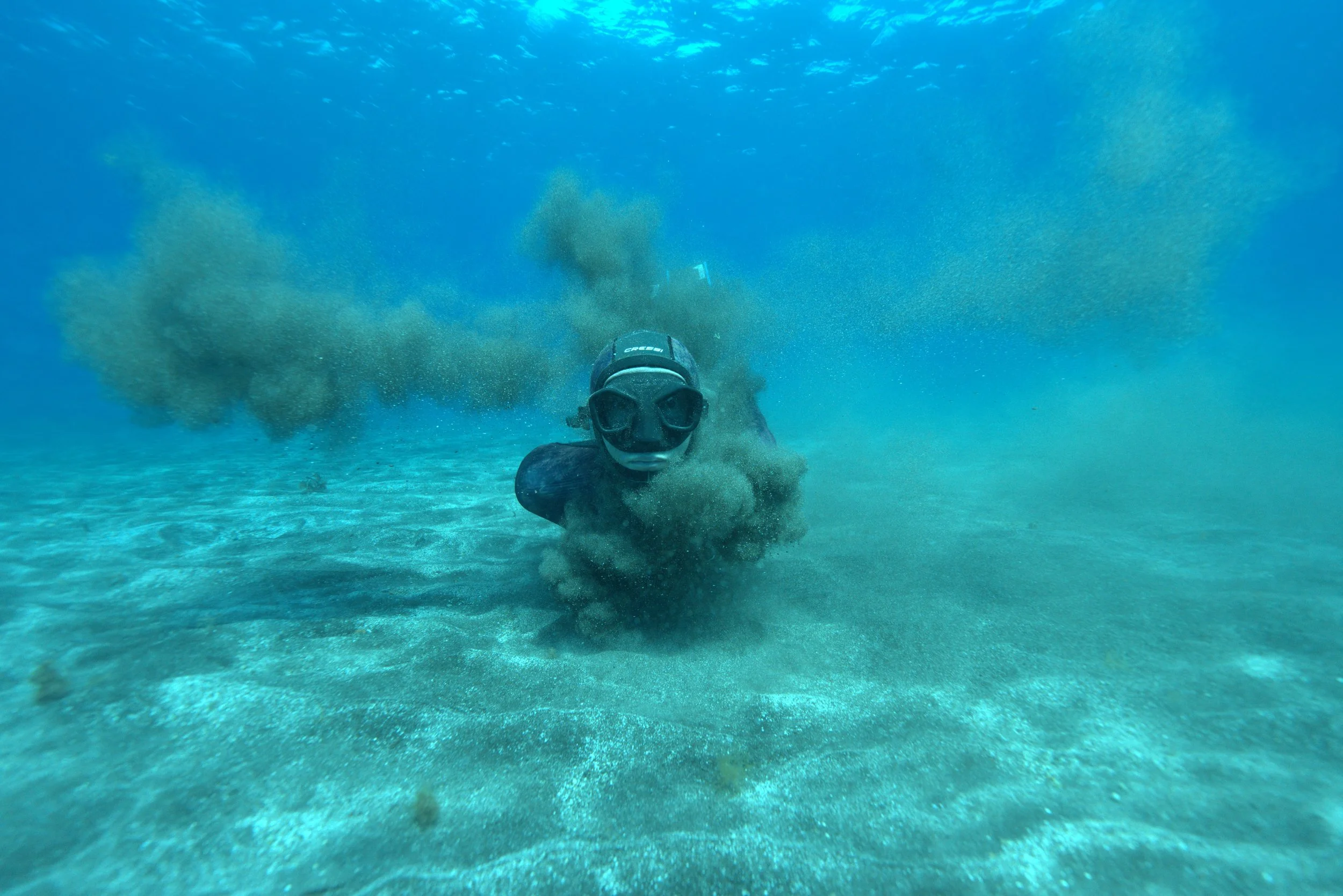 A freediver's face and mask on the bottom of the ocean, surrounded by a cloud of sand