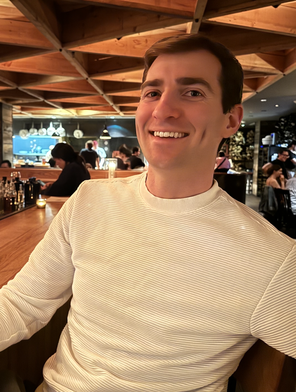 End-of-life doula. Doula Guild book club. A smiling man with short brown hair wearing a cream-colored ribbed long sleeve shirt, sitting at a wooden table in a lively restaurant with warm lighting, wooden ceiling, and group of people in background.