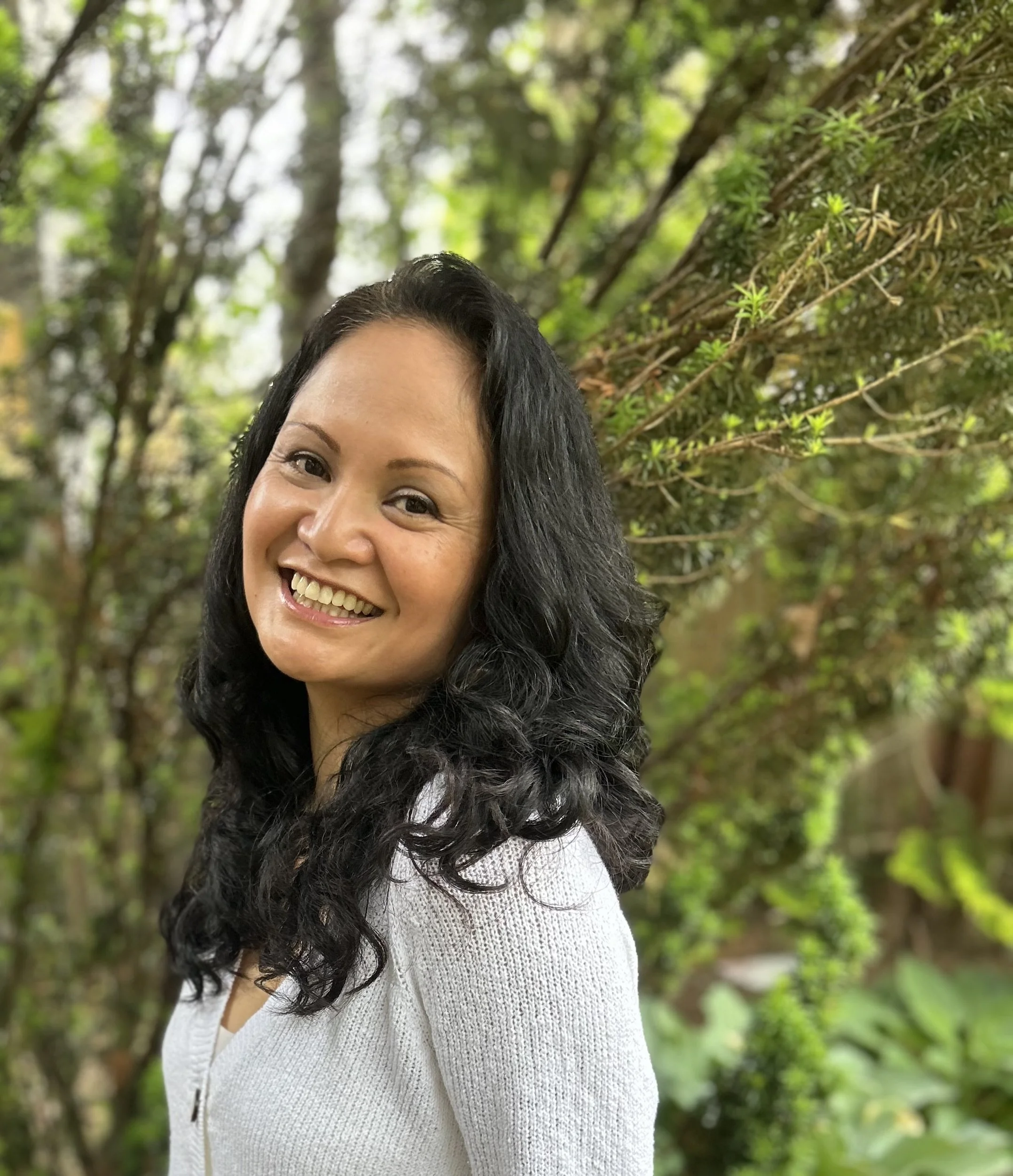 A woman with black, wavy hair smiling outdoors in a natural, green setting with trees and plants in the background.