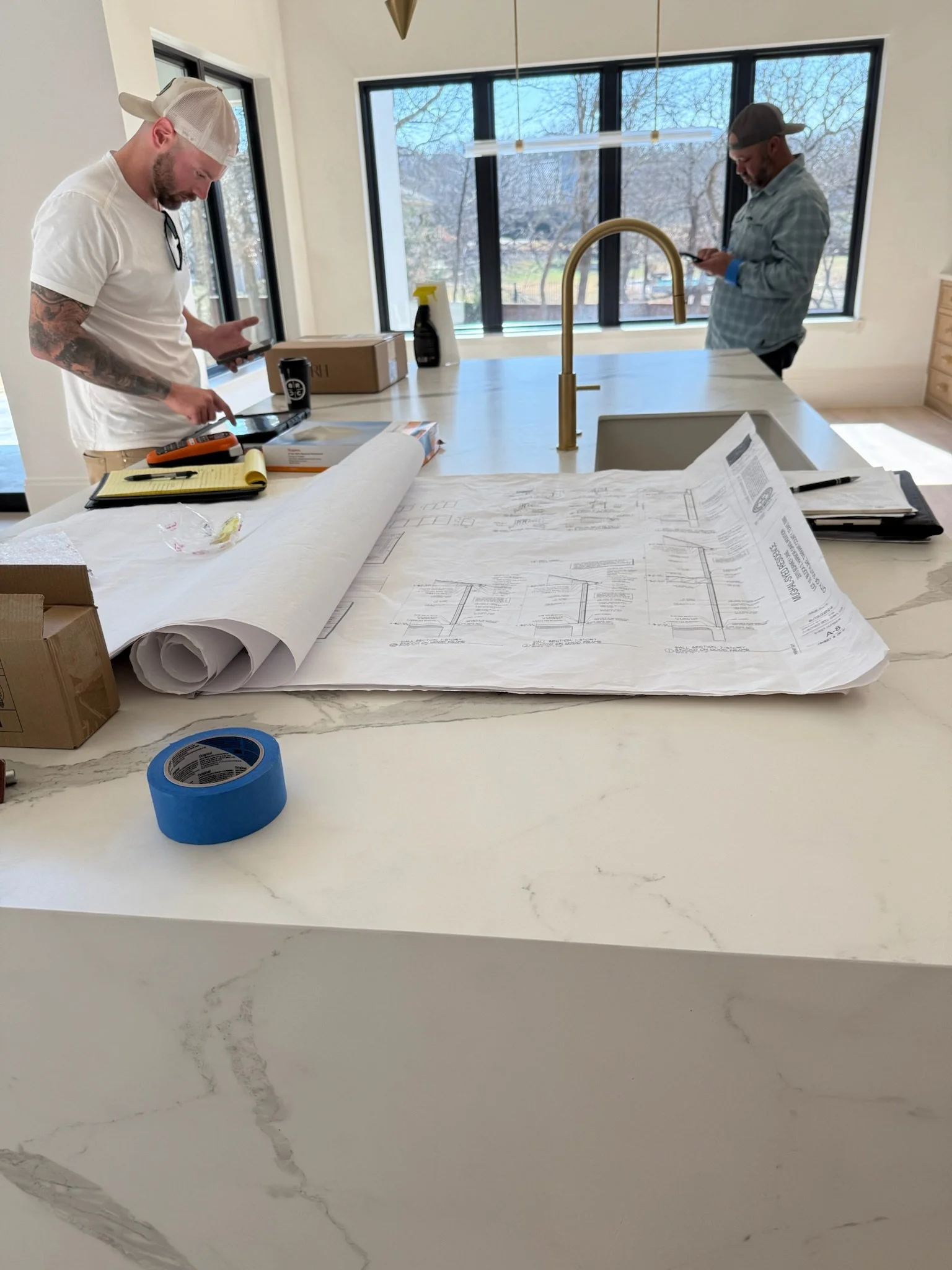 Two construction professionals reviewing architectural blueprints and wall section drawings on a marble waterfall kitchen island with brass faucet during a custom home walkthrough.