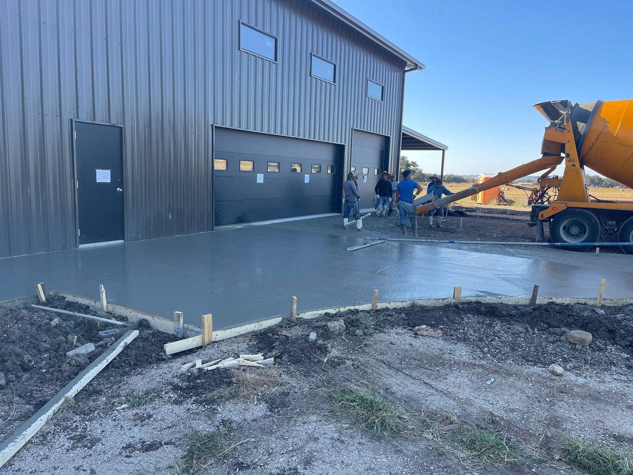 Concrete crew pouring and finishing a fresh concrete apron in front of a gray metal barndominium-style building with black roll-up garage doors in rural Texas.