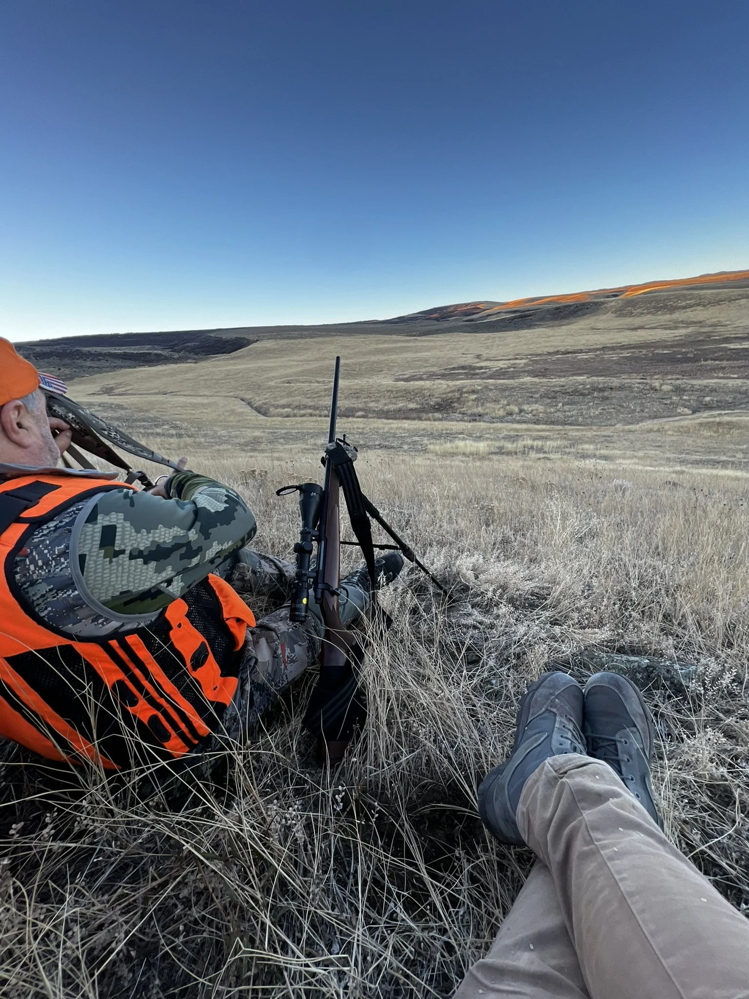 Two people seated in dry grass overlooking a hilly landscape, one with hunting gear and a rifle on a bipod, the other showing legs and boots. Clear blue sky overhead.