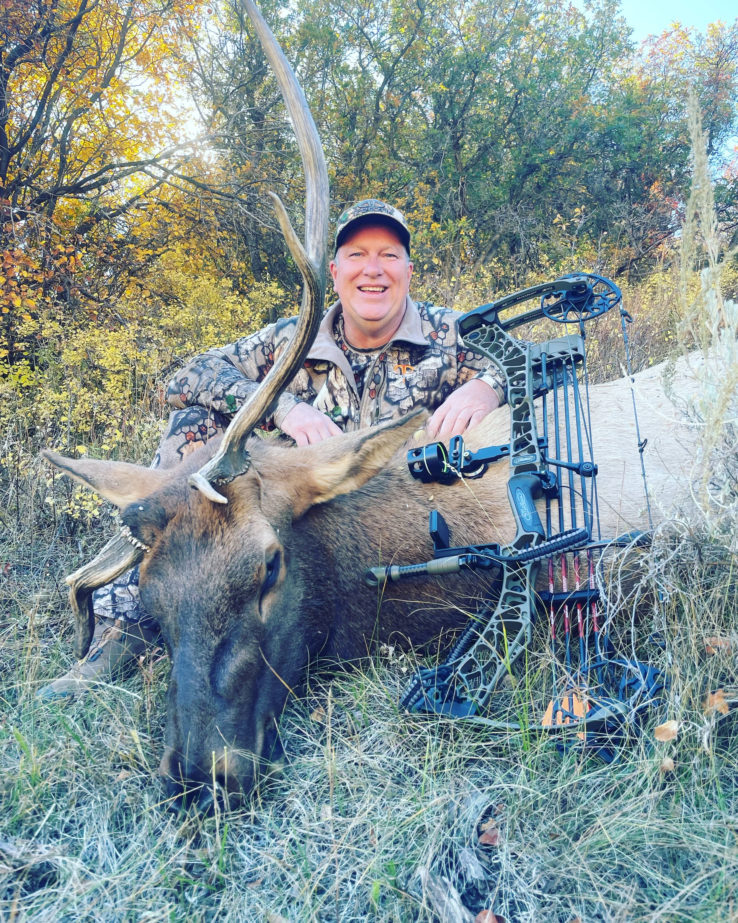 Hunter posing with a harvested elk and a compound bow in a forested area during fall.