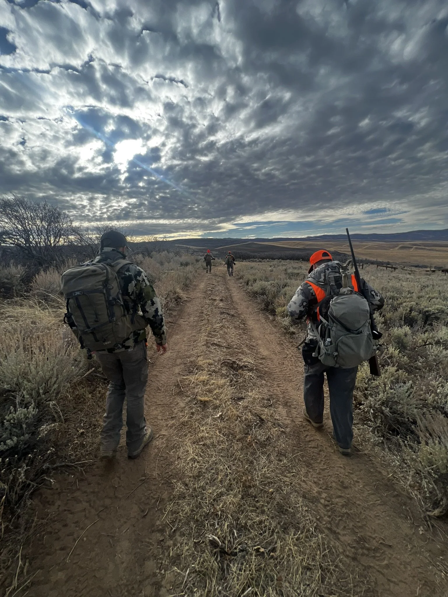 Group of hunters walking on a dirt path with backpacks and gear, under a cloudy sky in a dry, open landscape.