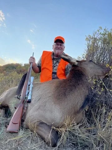 Hunter in orange vest with a rifle kneeling beside a hunted elk in a grassy area.