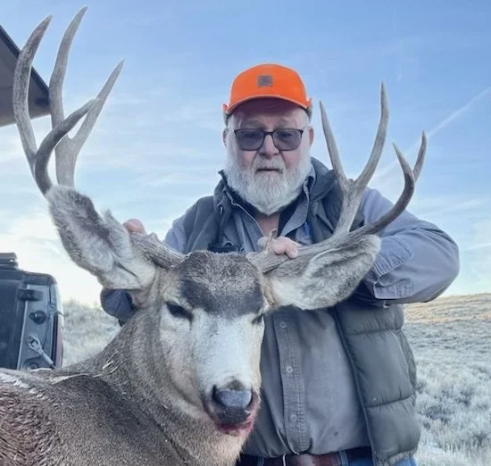 Man wearing orange cap standing next to a large deer with prominent antlers in an outdoor setting.