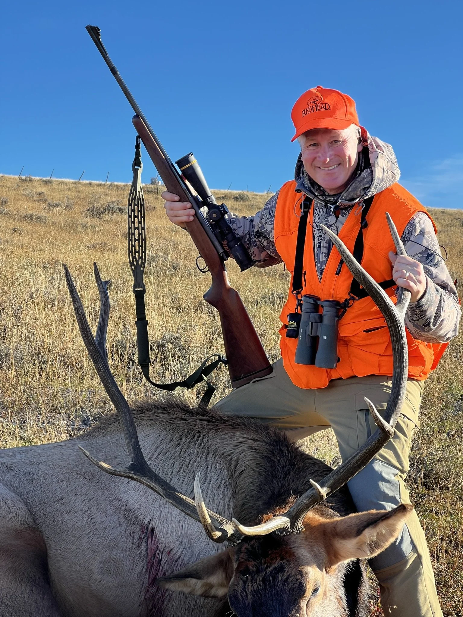 A smiling person in camouflage and bright orange hunting gear holds a rifle and poses with a hunted elk in a grassy field.