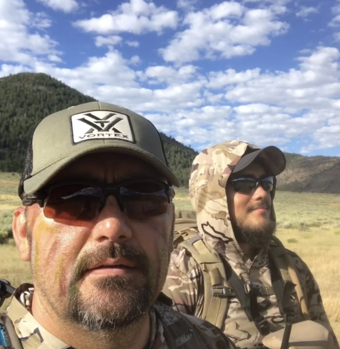 Two men wearing camouflage clothing and hats, standing outdoors in a scenic mountainous area with a blue sky and clouds in the background.