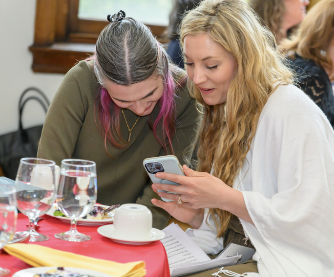 Darby Pistilli, LPC  and Michaela Uttley, LPC sitting at a table, smiling while looking at a phone together, representing friendship, voice notes, and the creation of Long Story Short, You Survive.