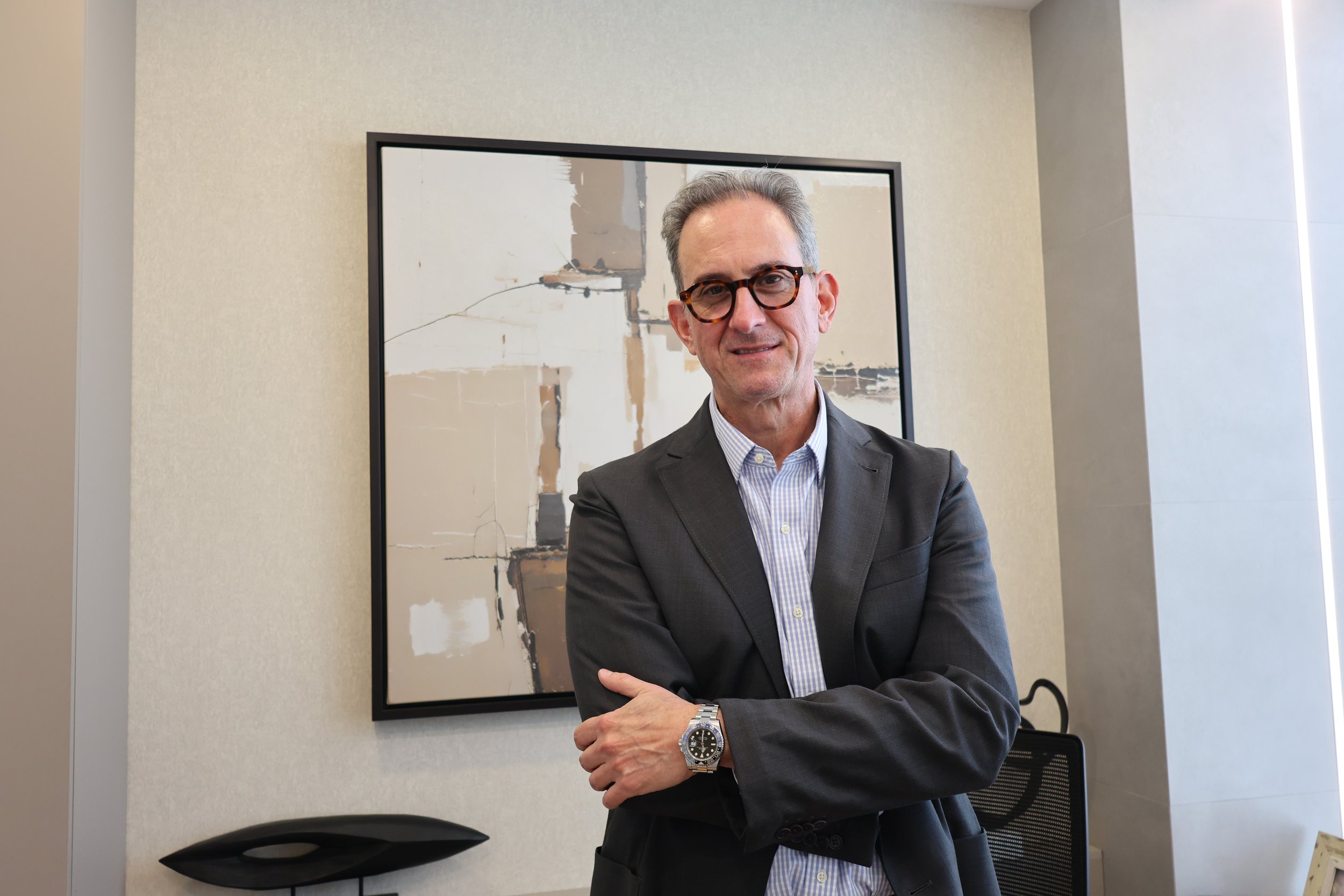 A middle-aged man with gray hair, wearing glasses and a dark suit, stands with arms crossed in an office, smiling at the camera, with abstract artwork behind him.