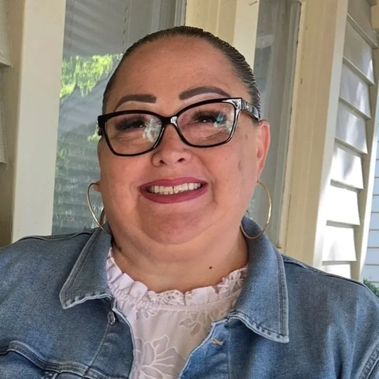A woman with glasses smiling in front of a window, wearing a denim jacket and a white top.