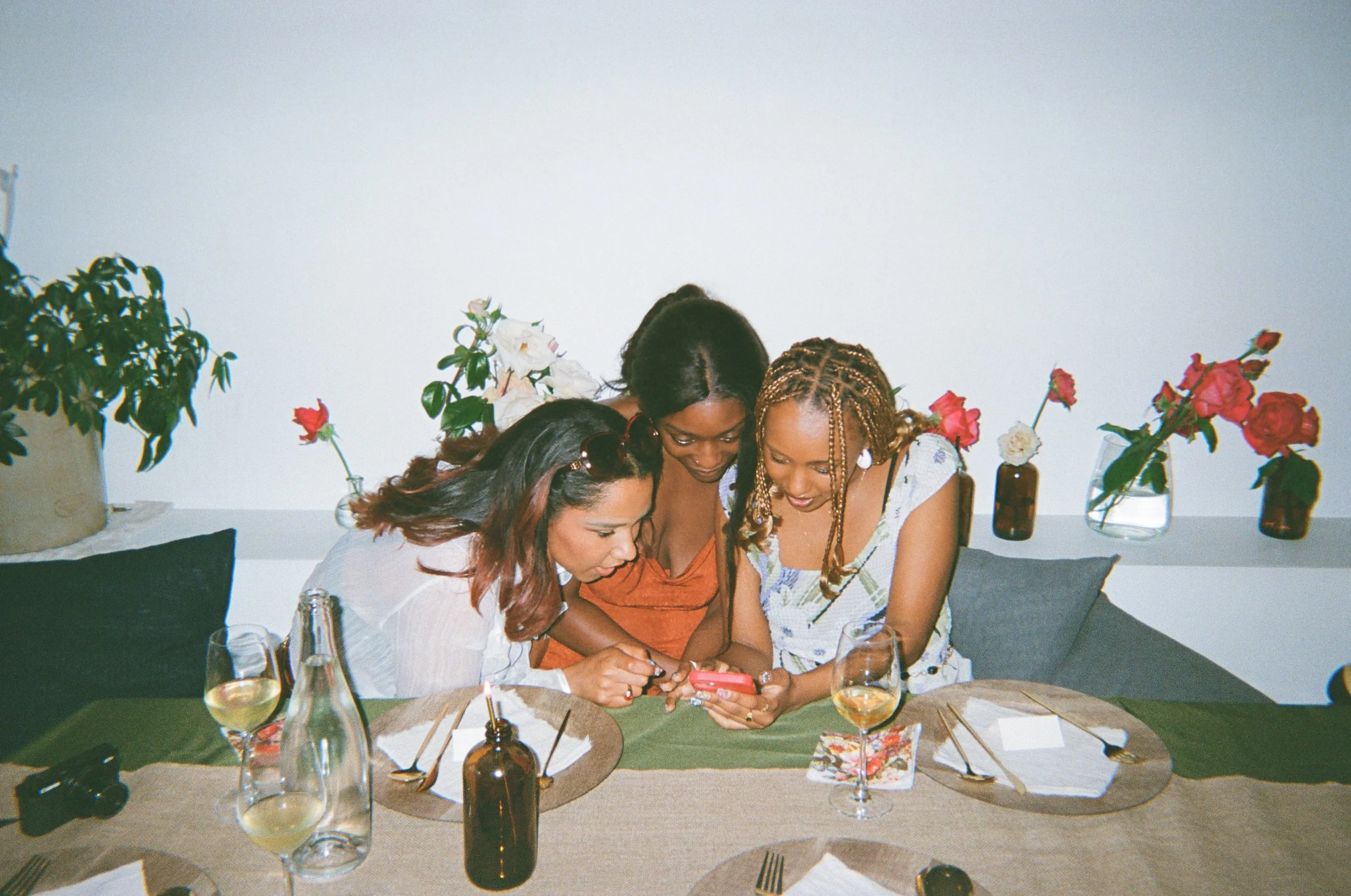Three black women are pictured sitting together at a booth behind a decorated rectangular dinner table. Behind them is a ledge with a row of vases holding one or two pink or white roses. No food has been served, and they each have a glass of bubbly o