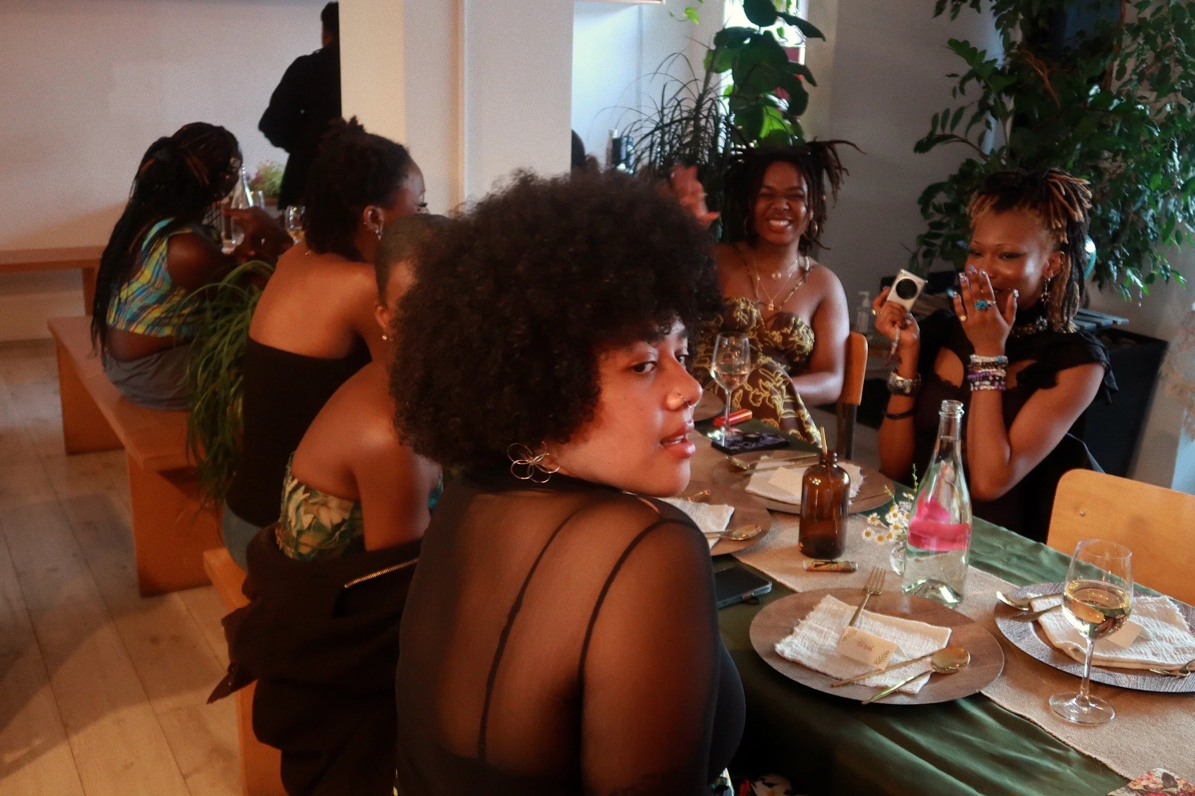 Friends are pictured sitting together on benches and laughing across a set and decorated rectangular dinner table. No food has been served. At the centre of the image is a light-skinned black woman with an afro looking over her shoulder, at the camer