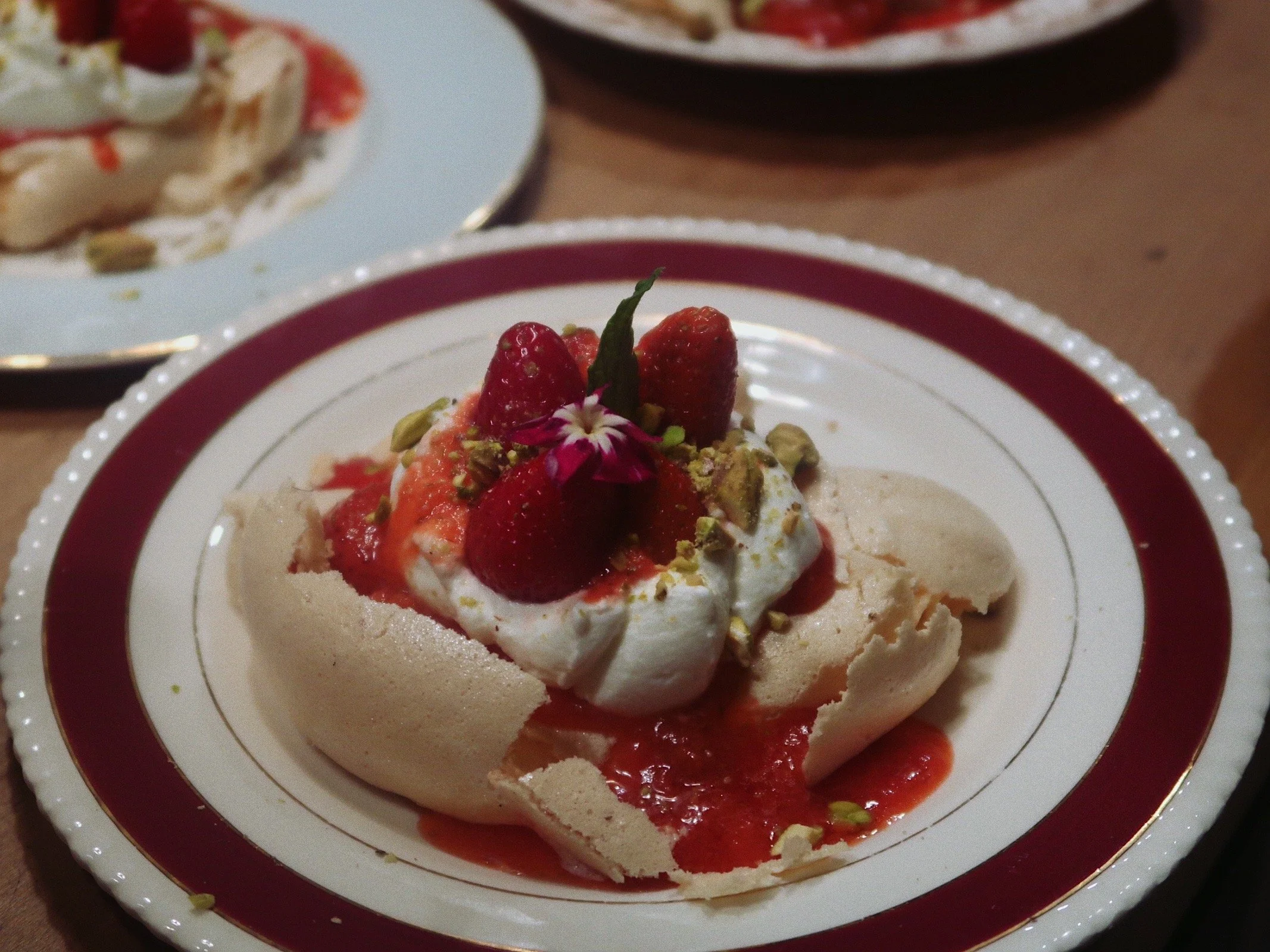 A close-up photograph of a plate of strawberry pavlova topped with whipped cream, strawberry sauce, pistachios, fresh strawberries, and an edible flower.