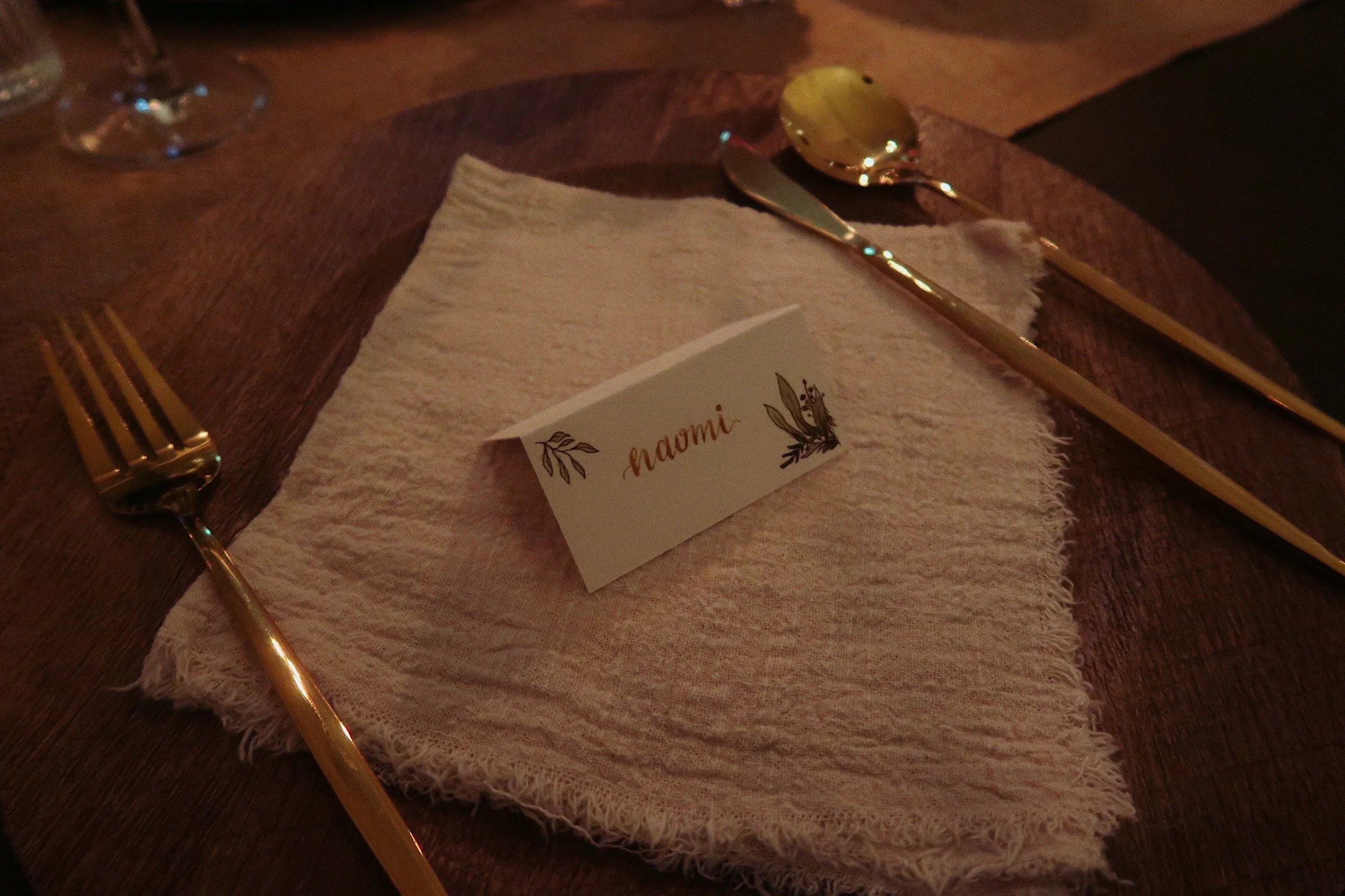 A zoomed-in photograph of a table setting. The image features a close-up of a faux-wood charger plate with gold cutlery, a cream-coloured napkin, and a place card featuring the name "Naomi" adorned with drawings of greenery. 
