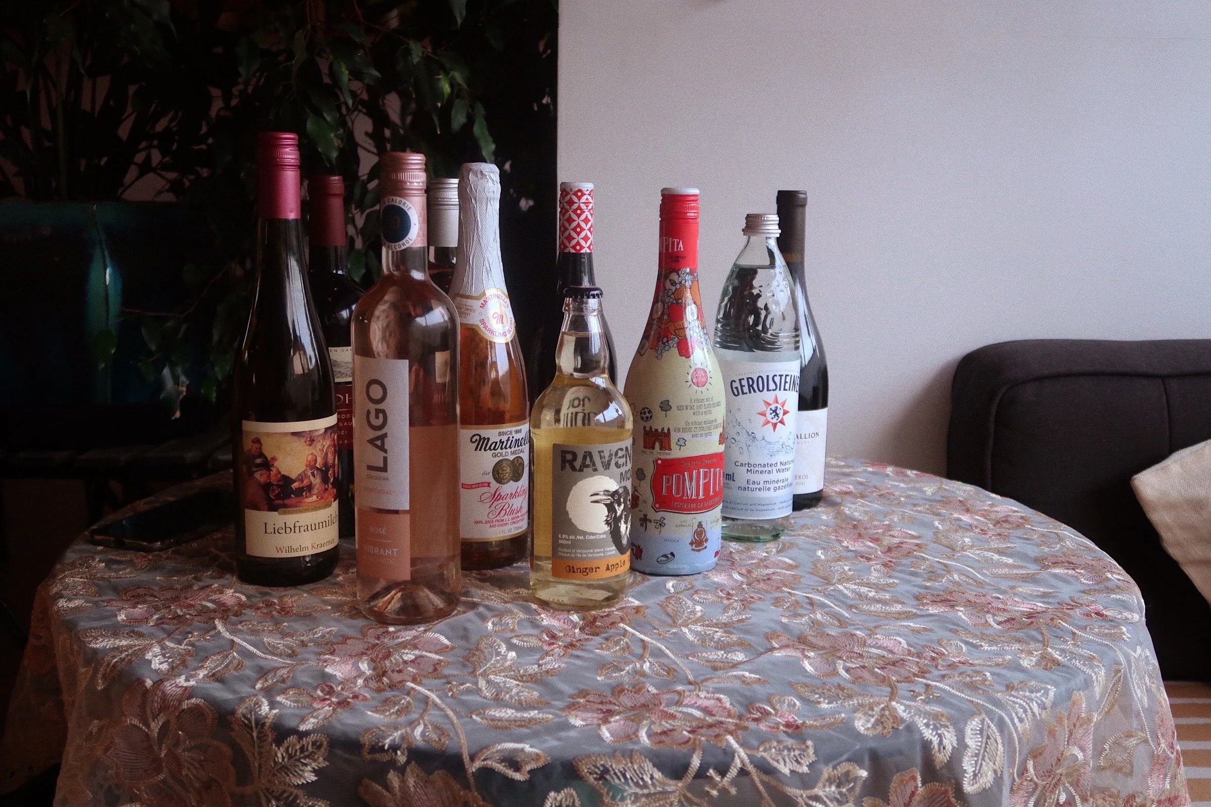 A circular table covered by a vintage lace tablecloth with a pink and cream floral pattern. The table holds eight bottles of different wines, a bottle of ginger apple cider, and a large bottle of sparkling water. All the bottles are made of glass.