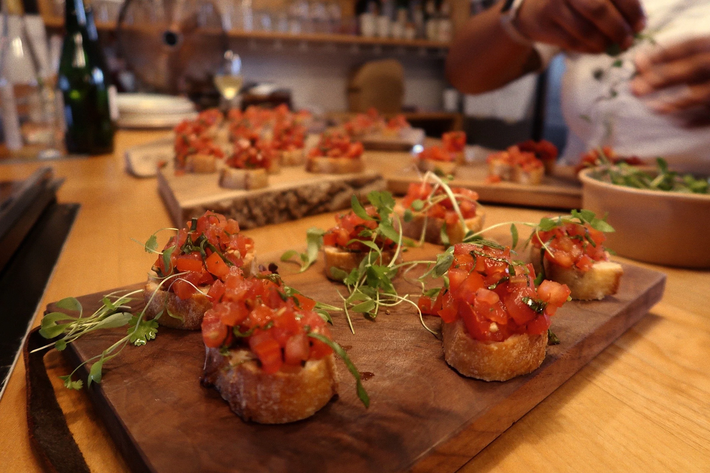 A photograph of assembled servings of balsamic bruschetta featuring baguette cuts topped with diced tomatoes, microgreens, and balsamic glaze. The bruschetta is being served on wooden boards. The chef's hands can be seen in the background, placing mi