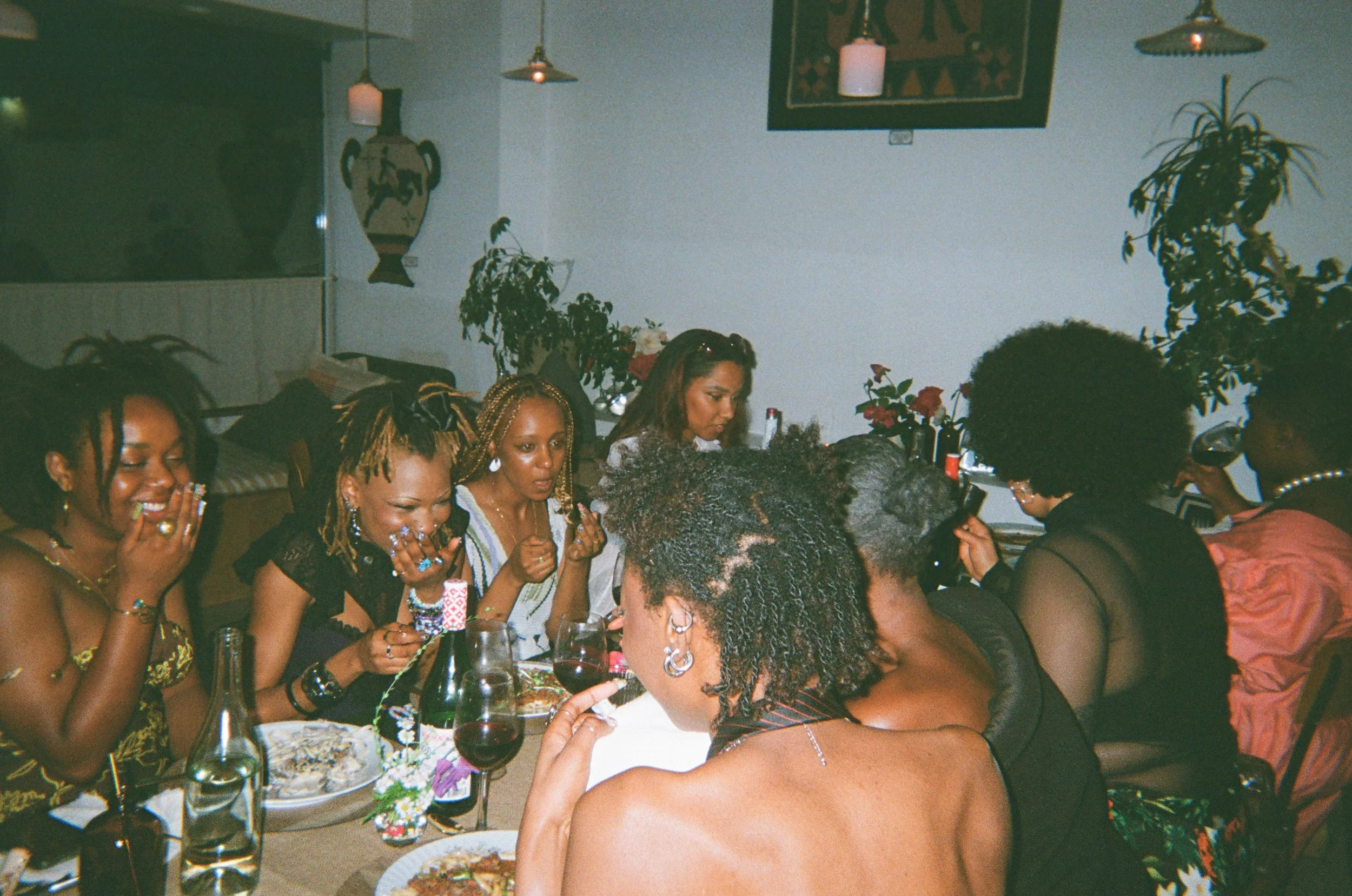 Friends are pictured sitting and laughing at a set and decorated dinner table. Pasta topped with a bolognese or mushroom sauce is in front of each person. At the centre of the image is a black woman with twists in her hair and her back to the camera.