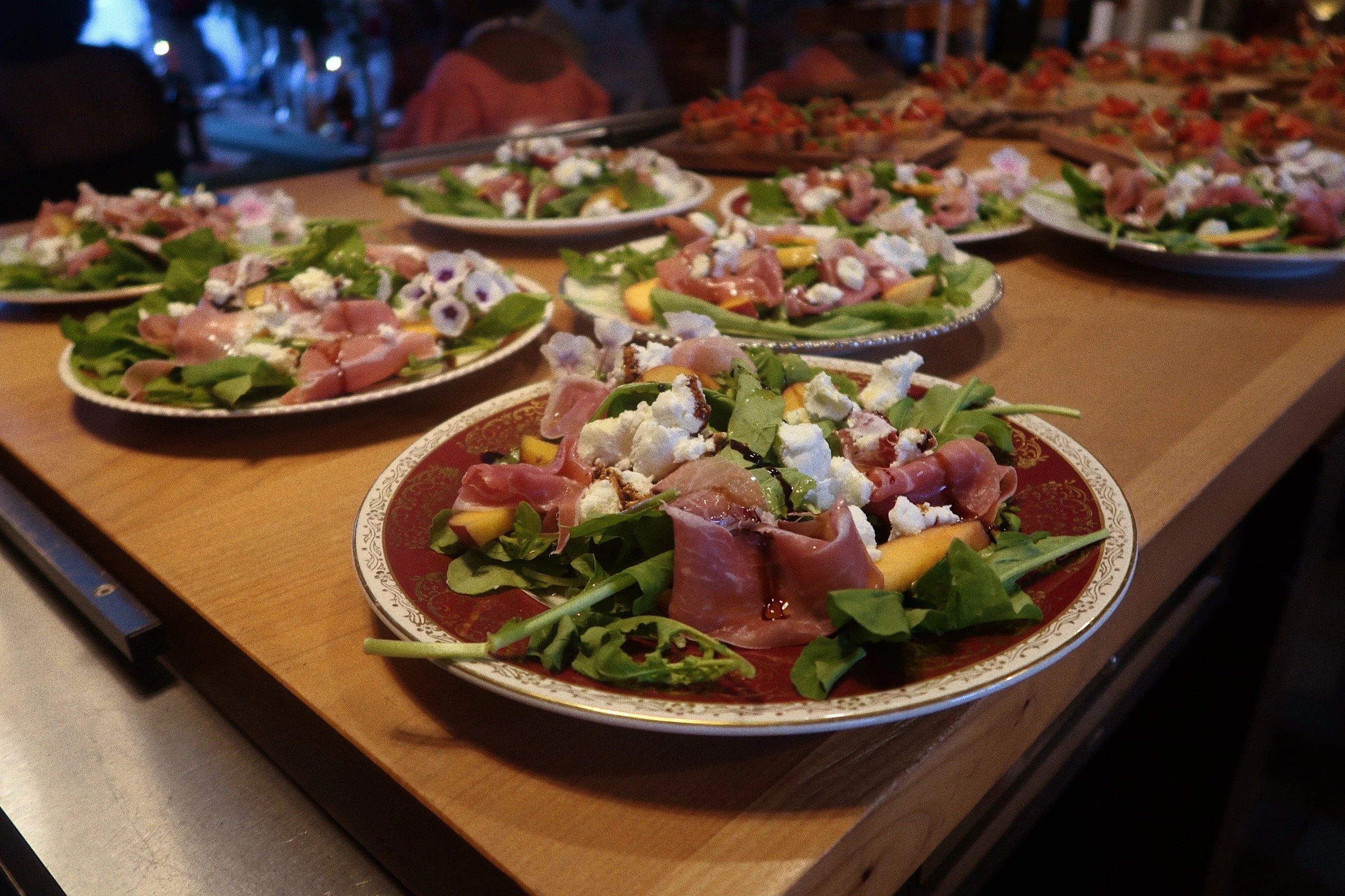 A photograph of a table featuring multiple plates of prosciutto, goat cheese and peach salad. The salad base is arugula, and each salad has been constructed on a vintage dish with a unique design.