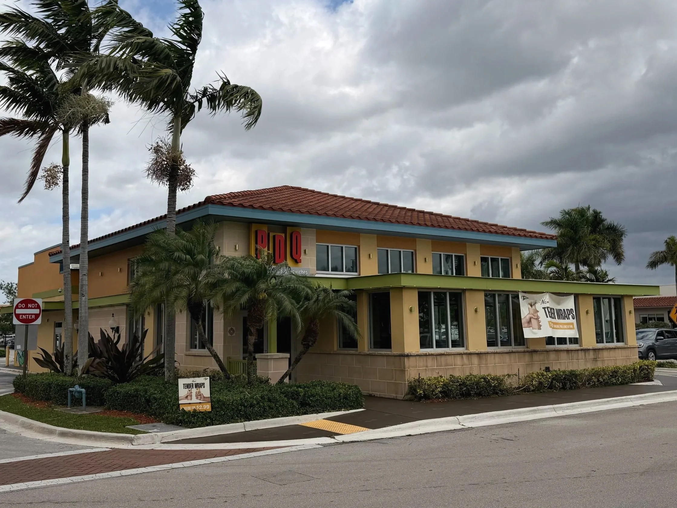 A commercial building with a red tile roof, palm trees in front, and a cloudy sky above. There are signs advertising tender wraps for $2.99.