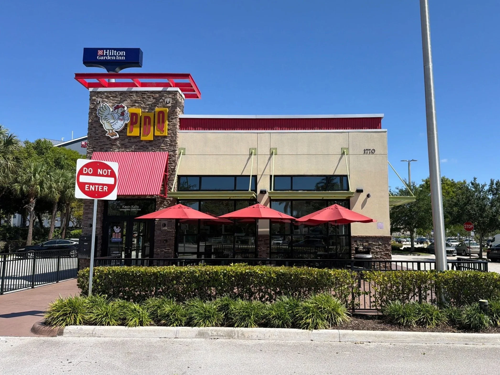 Exterior of a modern building with a sign for PDQ, a chicken restaurant, featuring a rooster logo. There are red umbrellas outside and a "Do Not Enter" sign on the left. Tall palm trees are visible on the left side, and the sky is clear and blue.