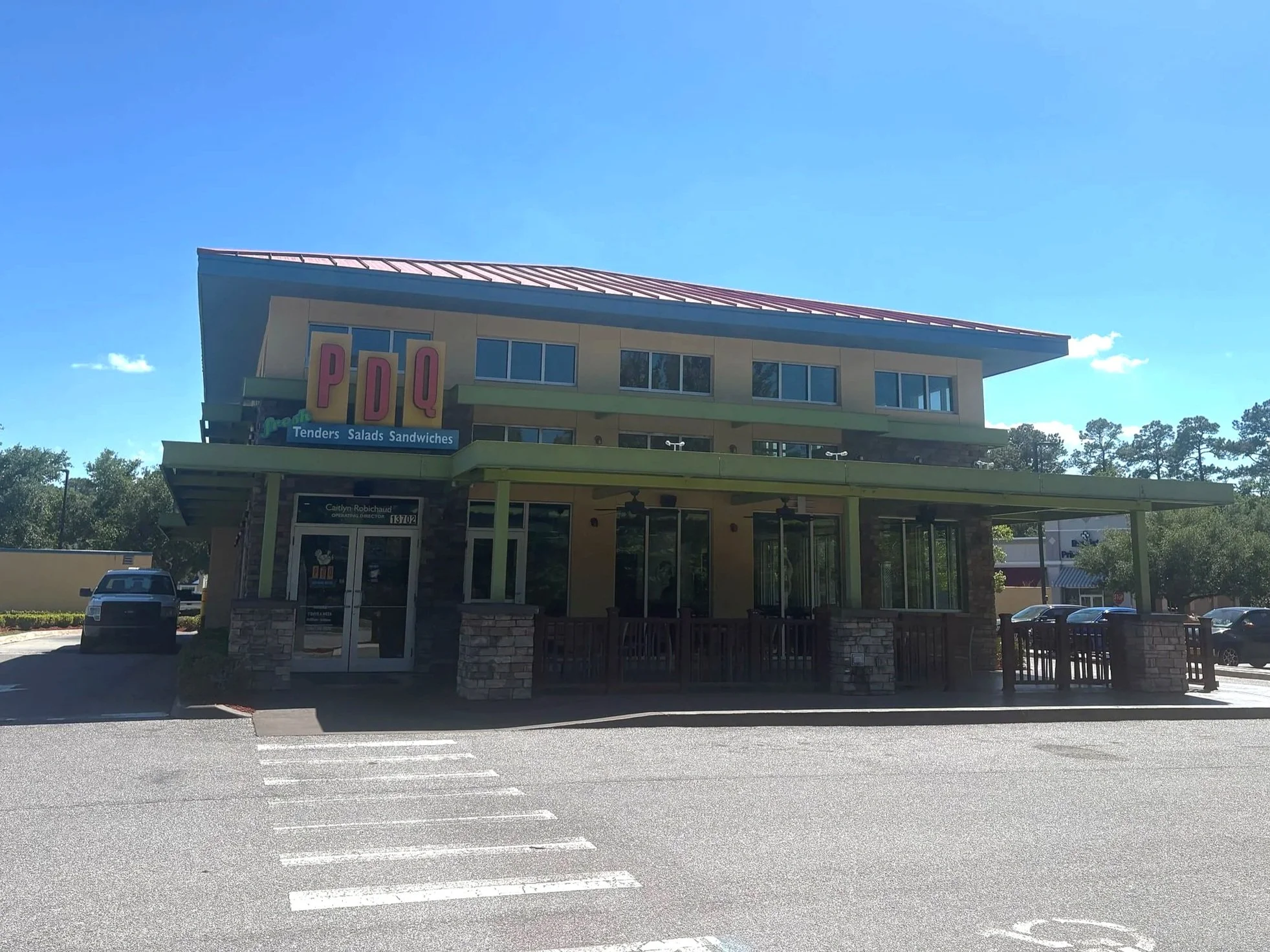 A two-story restaurant building with a green roof and beige exterior, featuring large windows. A prominent sign reads 'PDQ' and advertises tenders, salads, and sandwiches. There is a parking lot in front with a few cars and a clear blue sky overhead.