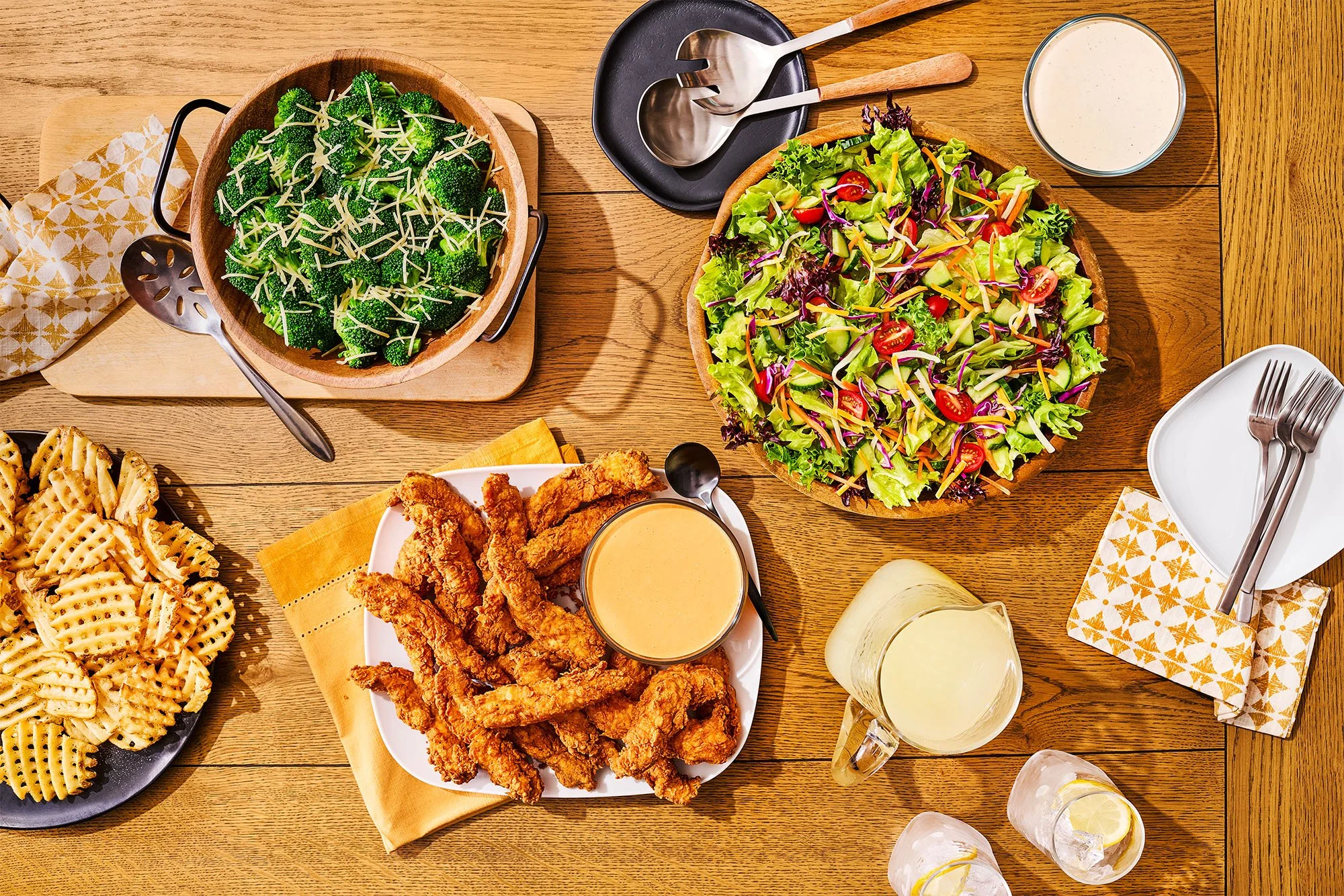 A catering spread with chicken tenders, waffle fries, salad, broccoli, lemonade, and sauces.