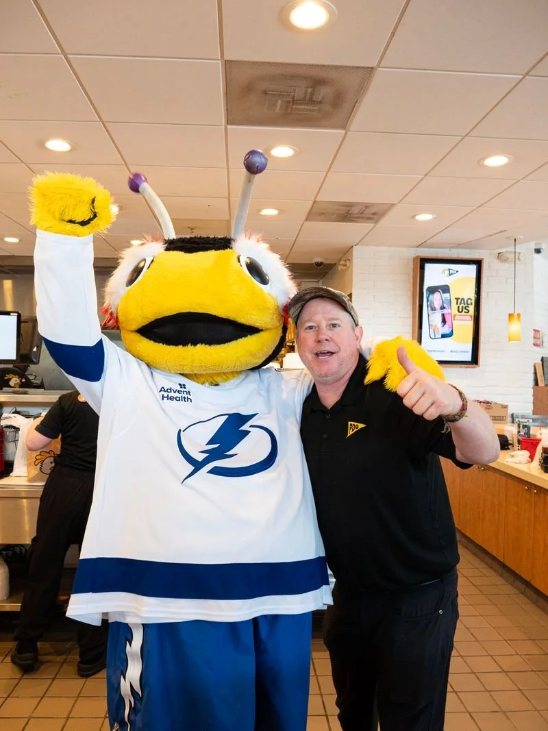A person in a Tampa Bay Lightning hockey jersey standing next to a large mascot resembling a yellow wasp with antennae, inside a fast-food restaurant or cafe, making a thumbs-up gesture.