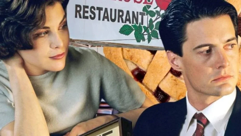 A woman with short dark hair and a man with dark hair and a suit sit in front of a restaurant sign, with a pizza behind them.