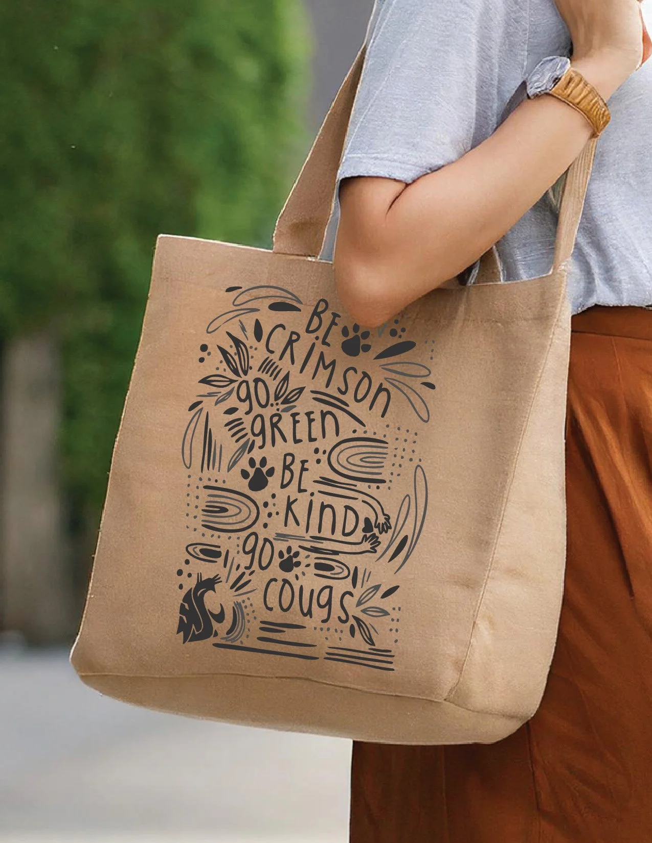 A person carrying a brown tote bag with black printed text that encourages kindness and respect for animals, including phrases like 'Be a crimport,' 'Go green,' 'Be kind,' and 'Go cougars.'