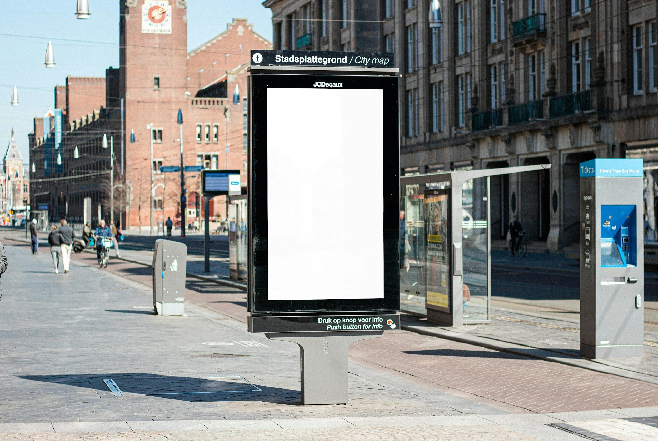 City street with digital billboard, tram ticket kiosk, and a few pedestrians walking on sidewalk.