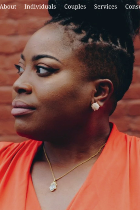 Profile of an African American woman with styled dreadlocks, wearing pearl earrings, a gold necklace with a pendant, and an orange top, standing against a brick wall.